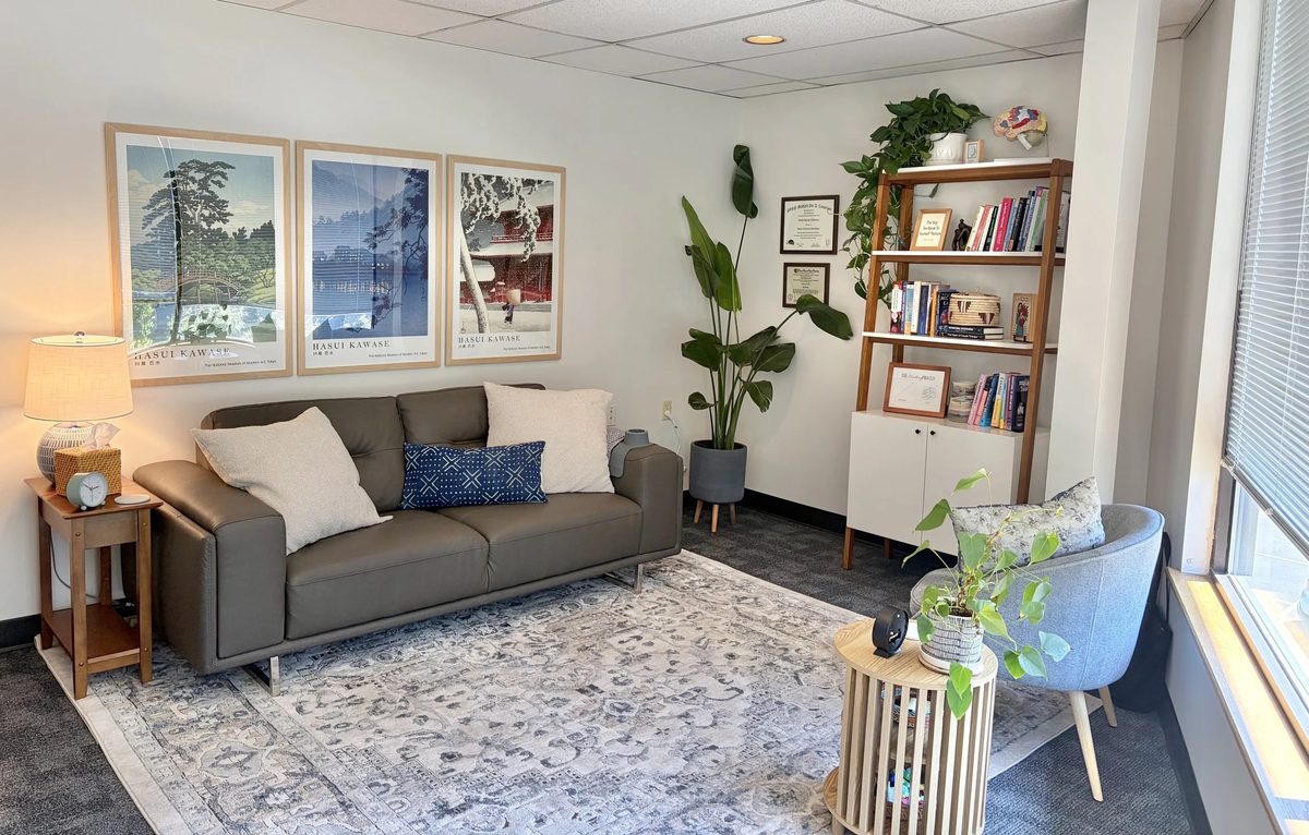 A softly lit room with a sofa, an armchair, plants, a bookshelf, and framed prints